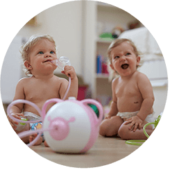 Two happy baby girls sitting on the floor with the Nosiboo Pro Electric Nasal Aspirator