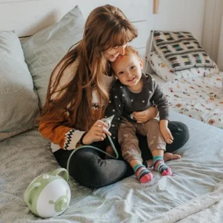 A mom sitting with her happy daughter on a bed and holding a Nosiboo Pro Electric Nasal Aspirator