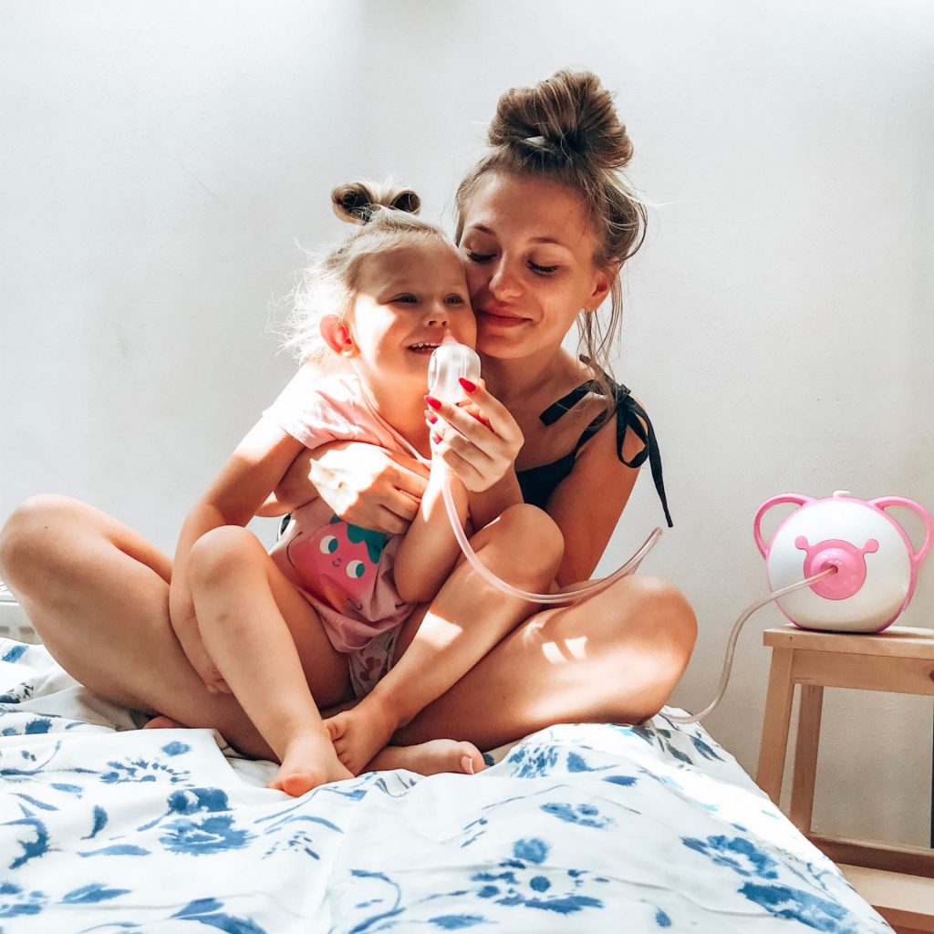 Mamma sorridente e sua figlia sedute su un letto e usano l'aspiratore nasale elettrico Nosiboo Pro